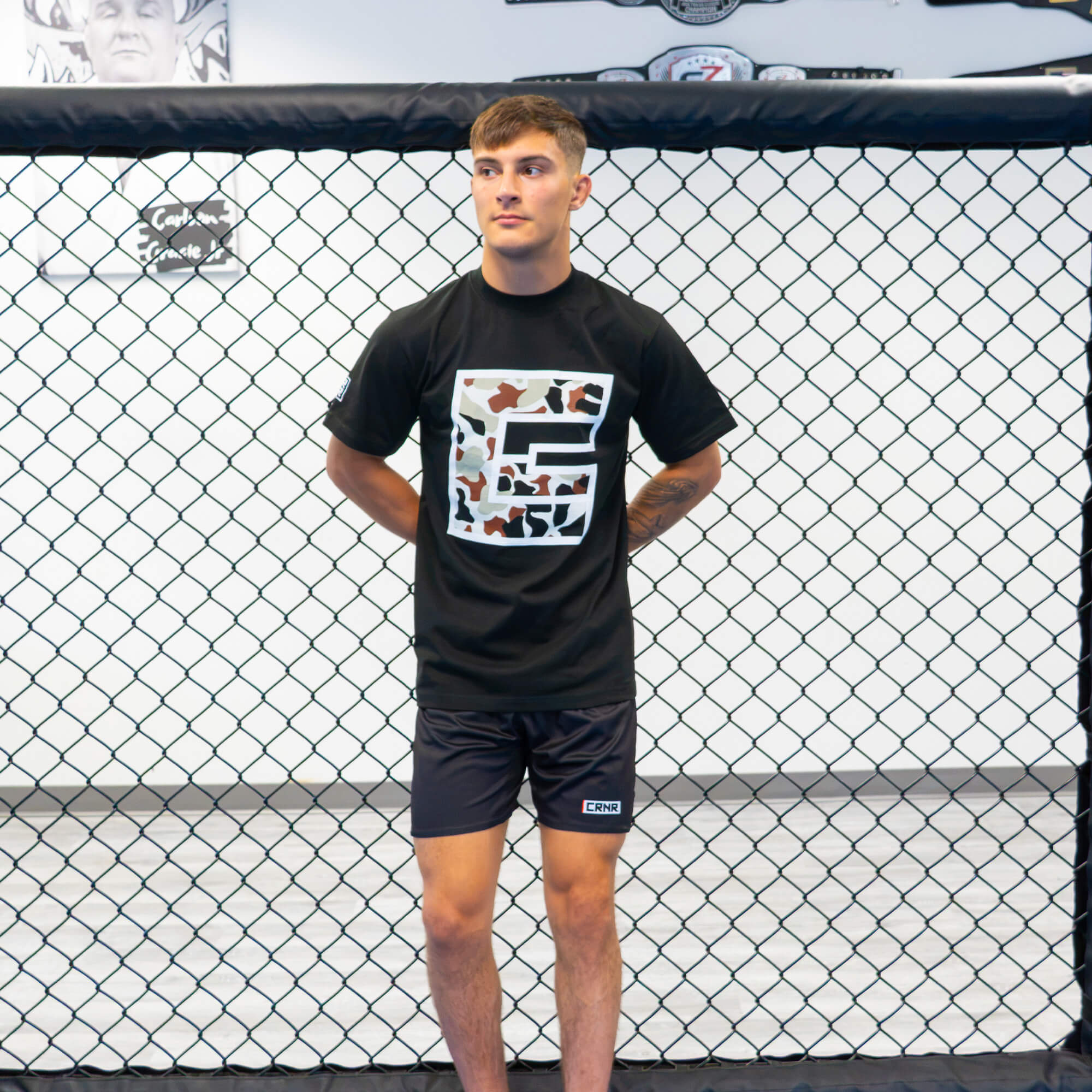 Full-length portrait of a male fighter standing inside an MMA cage with his hands behind his back, wearing the Camo Icon T-Shirt and matching black training shorts.