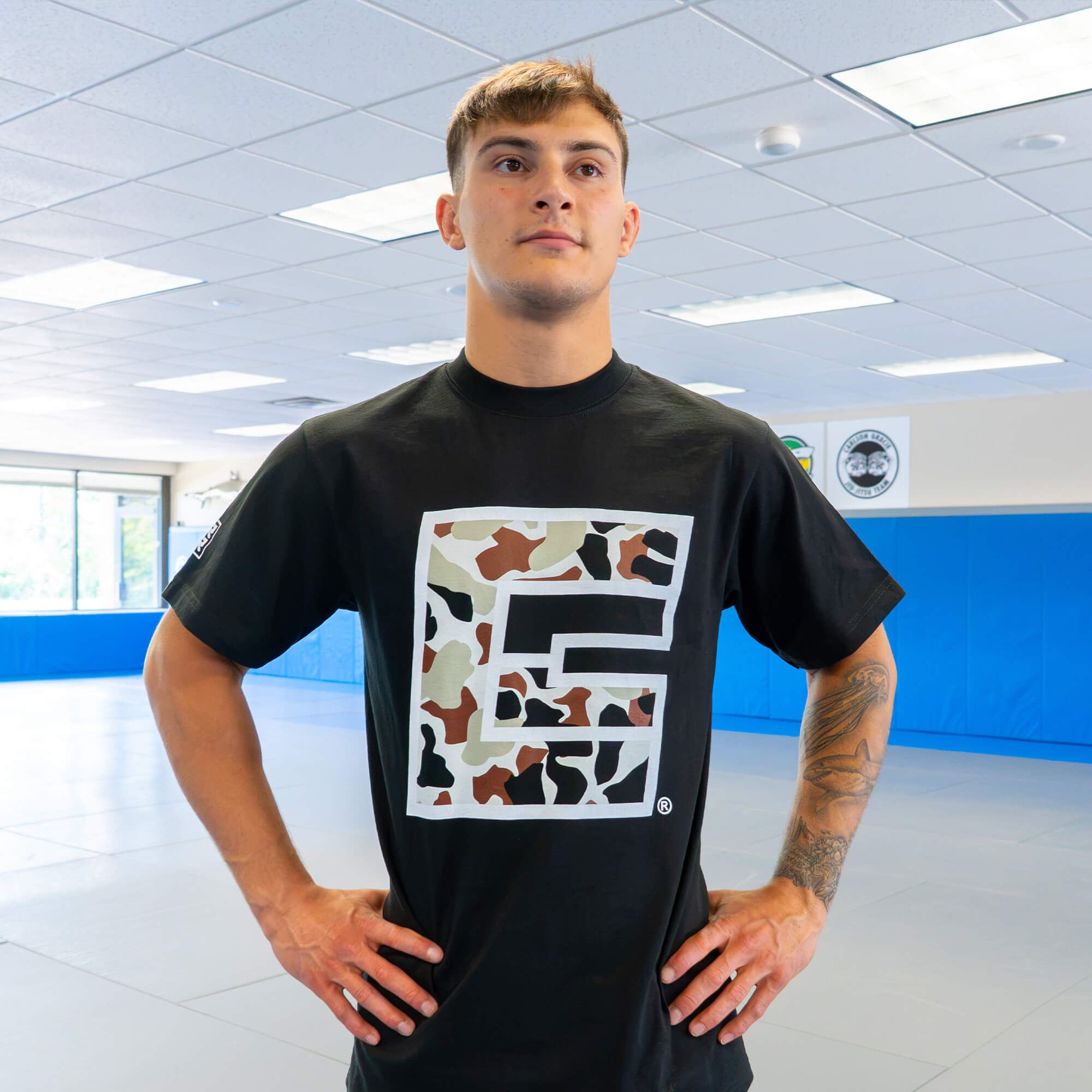 A young male athlete with a forearm tattoo standing hands-on-hips inside a gym, wearing the Camo Icon T-Shirt with a large camo-patterned square icon on the front.