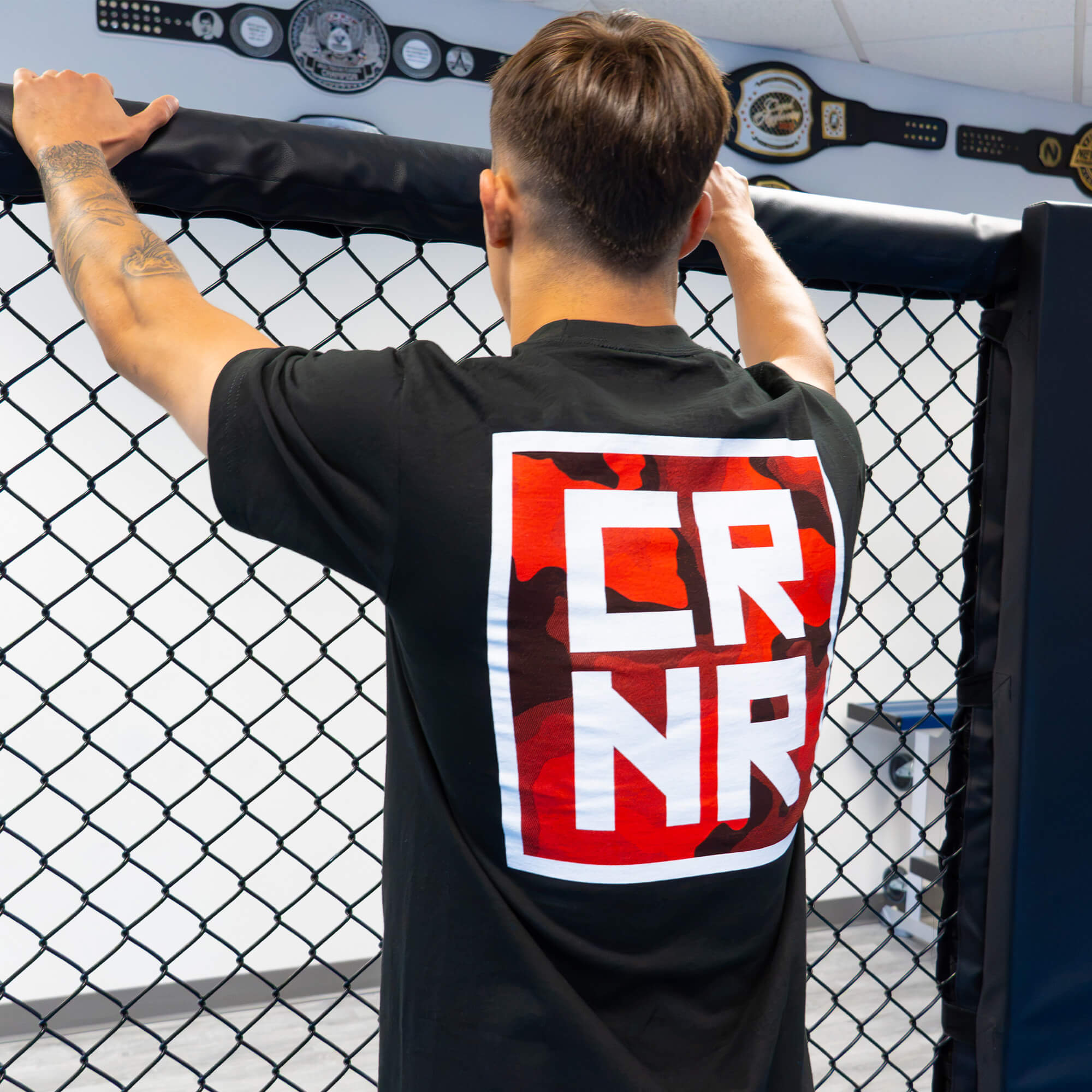Rear view of a male athlete standing at an MMA cage fence, wearing  with a red and white square logo on the chest featuring a large red and white stacked "CRNR" graphic on the back.