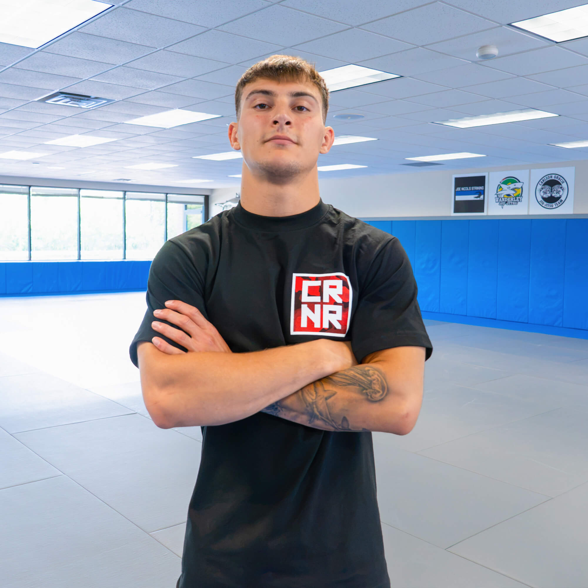A young male fighter standing with crossed arms inside a martial arts gym, wearing the Cinder Topo Stacked T-Shirt with a red and white square logo on the chest.