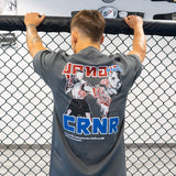 Rear view of a male athlete standing at an MMA cage fence, wearing the Golden Era Thai Boxing charcoal grey crewneck t-shirt featuring a large graphic of Muay Thai fighters and blue "CRNR" text on the back.