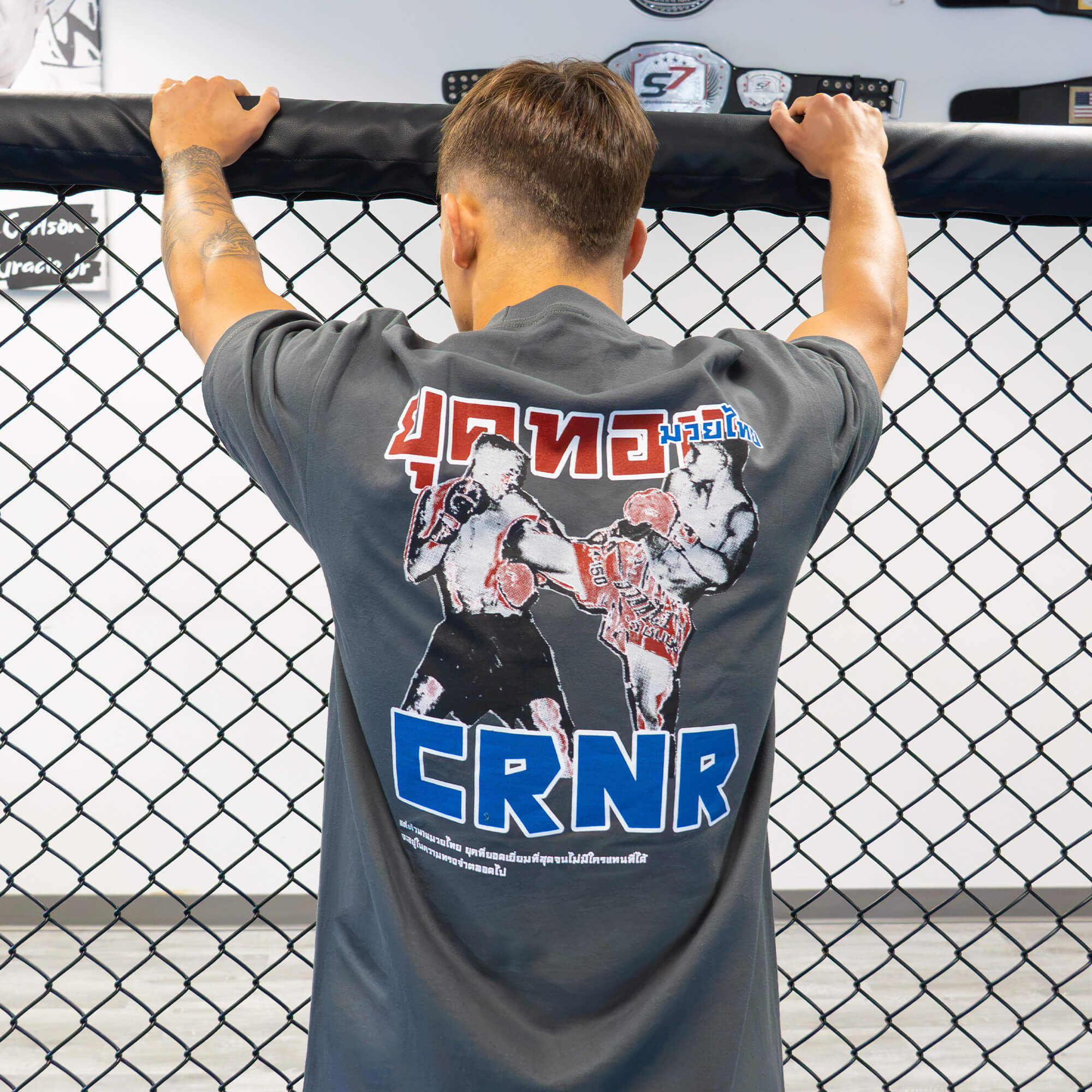 Rear view of a male athlete standing at an MMA cage fence, wearing the Golden Era Thai Boxing charcoal grey crewneck t-shirt featuring a large graphic of Muay Thai fighters and blue "CRNR" text on the back.