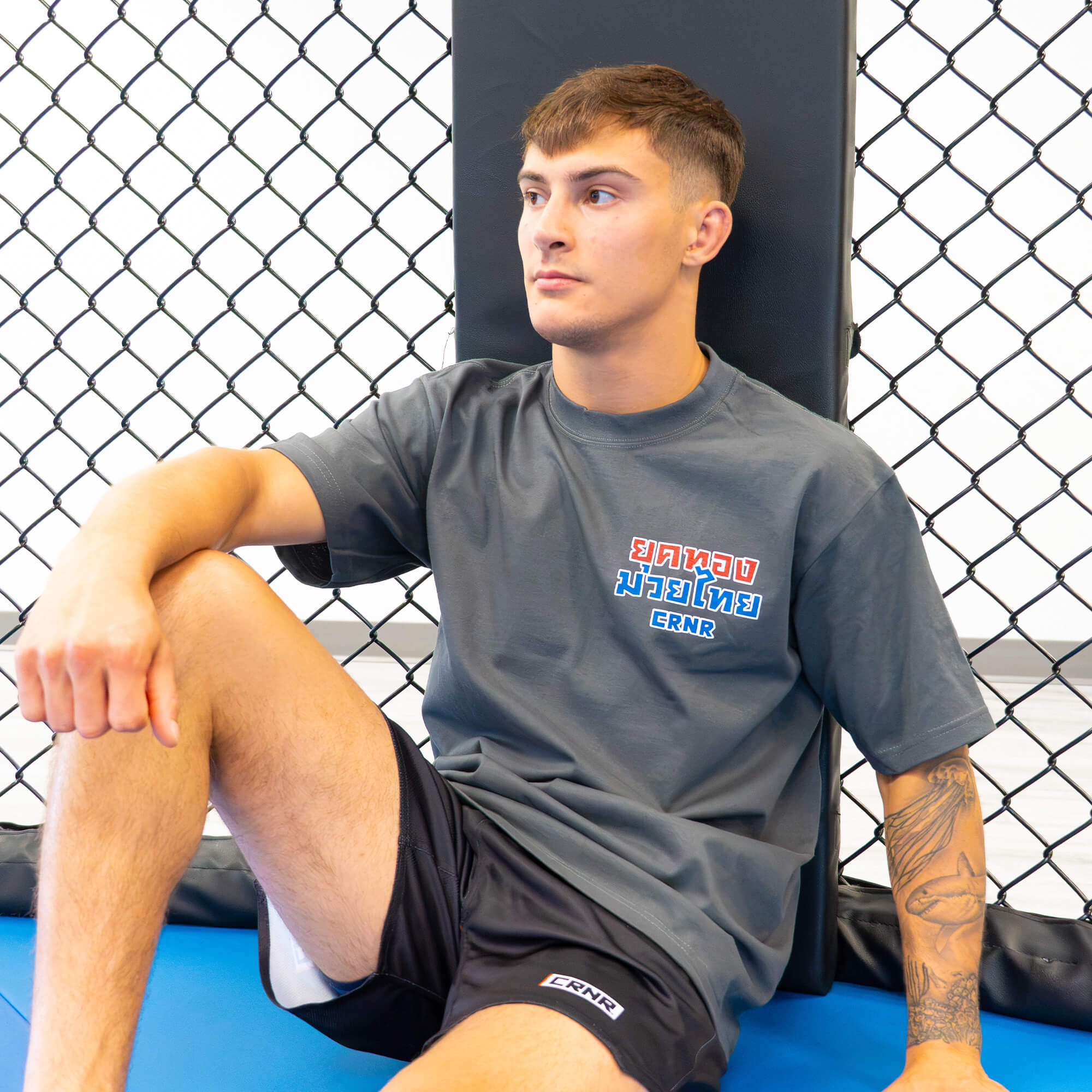A young male fighter with a forearm tattoo sits relaxed against an MMA cage corner pad, wearing the Golden Era Thai Boxing charcoal grey crewneck t-shirt with red and blue Thai script on the chest.