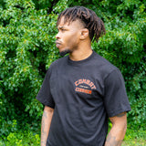 Side profile view of a man with dreadlocks looking to the left, standing in front of lush green bushes. He is wearing a black Combat Corner t-shirt that features an orange and red block-letter logo on the left chest reading "Combat Fight Shop Corner"