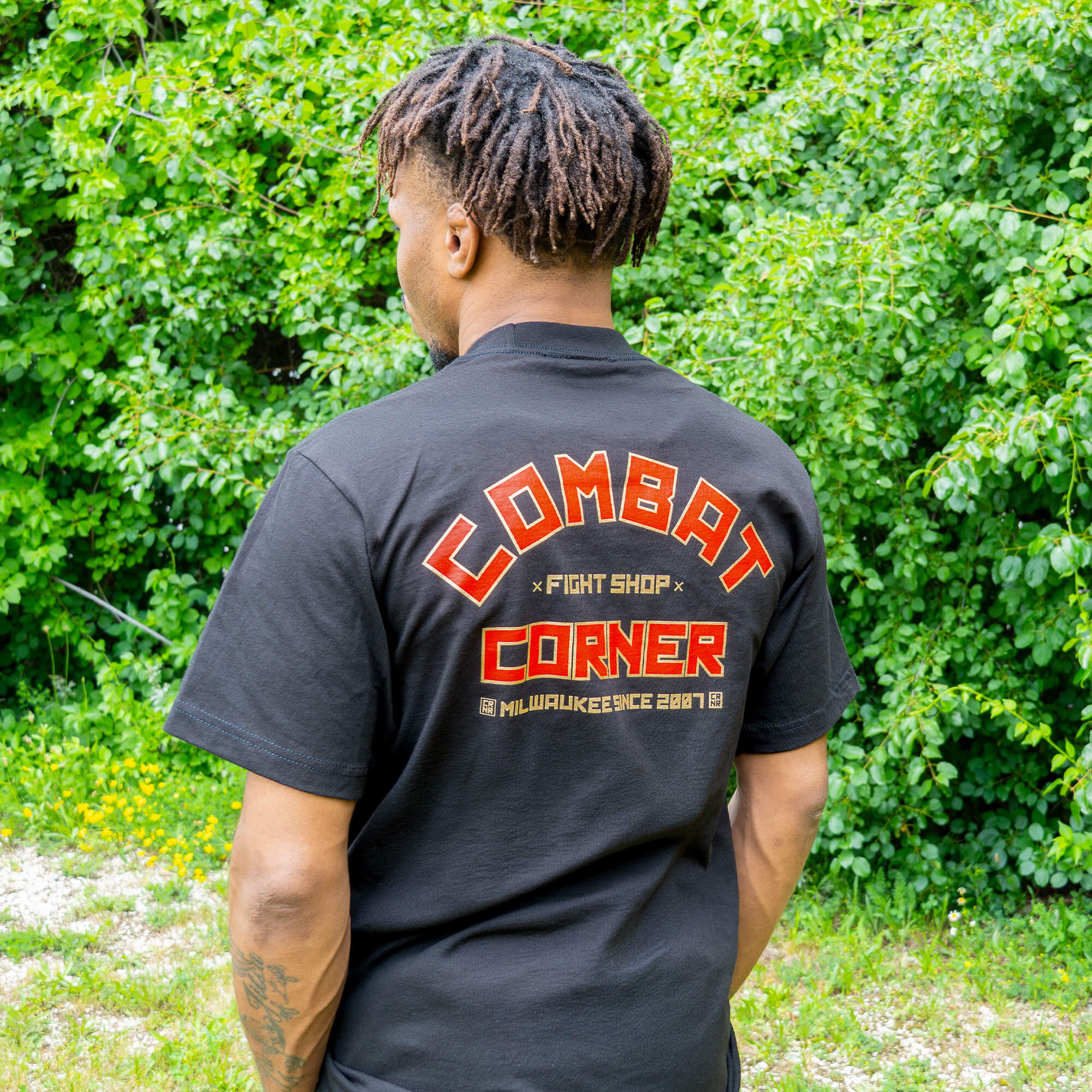 Rear view of a man looking away, showcasing the large graphic on the back of a black Combat Corner t-shirt. The vibrant orange and red design features arched block letters that read "Combat Fight Shop Corner" above the text "Milwaukee Since 2007"