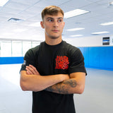 A male athlete standing with arms crossed in a training facility, modeling the front of the black Combat Corner HWD Ancient t-shirt to show its chest graphic and sleeve length.
