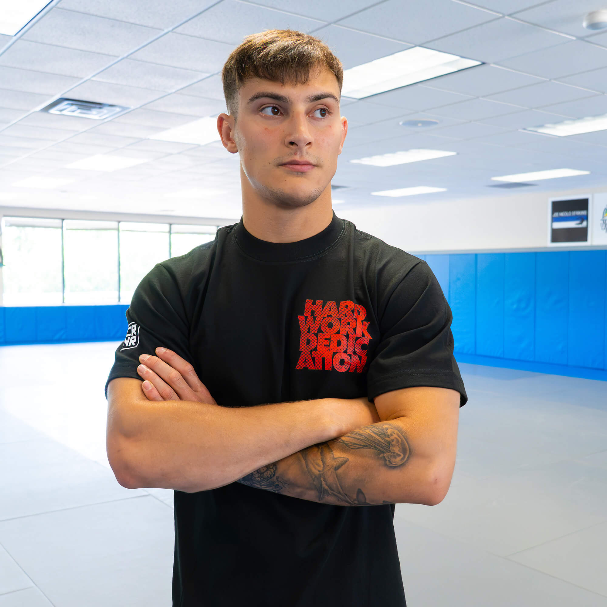 A male athlete standing with arms crossed in a training facility, modeling the front of the black Combat Corner HWD Ancient t-shirt to show its chest graphic and sleeve length.