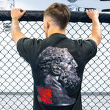 Rear view of a male athlete holding onto the top of an MMA cage, showcasing the large classical statue and red typography graphic on the back of the black Combat Corner t-shirt.
