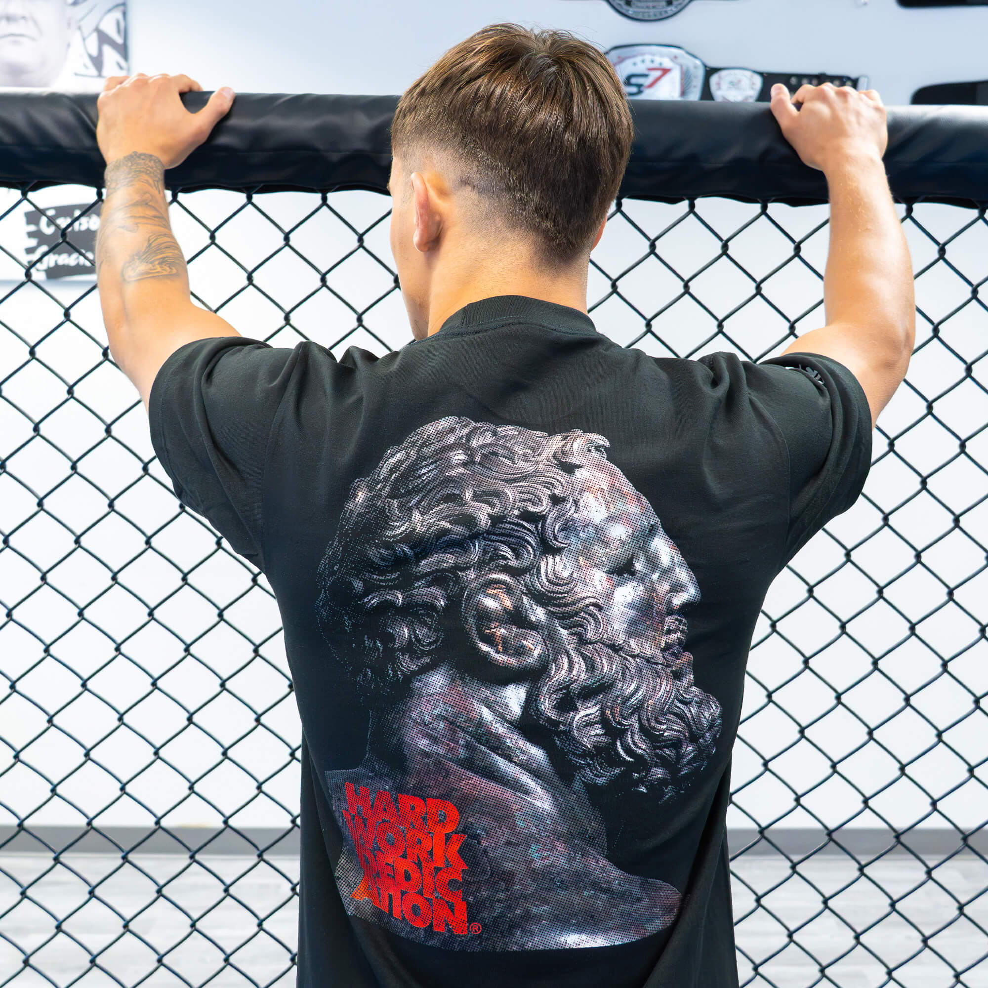 Rear view of a male athlete holding onto the top of an MMA cage, showcasing the large classical statue and red typography graphic on the back of the black Combat Corner t-shirt.