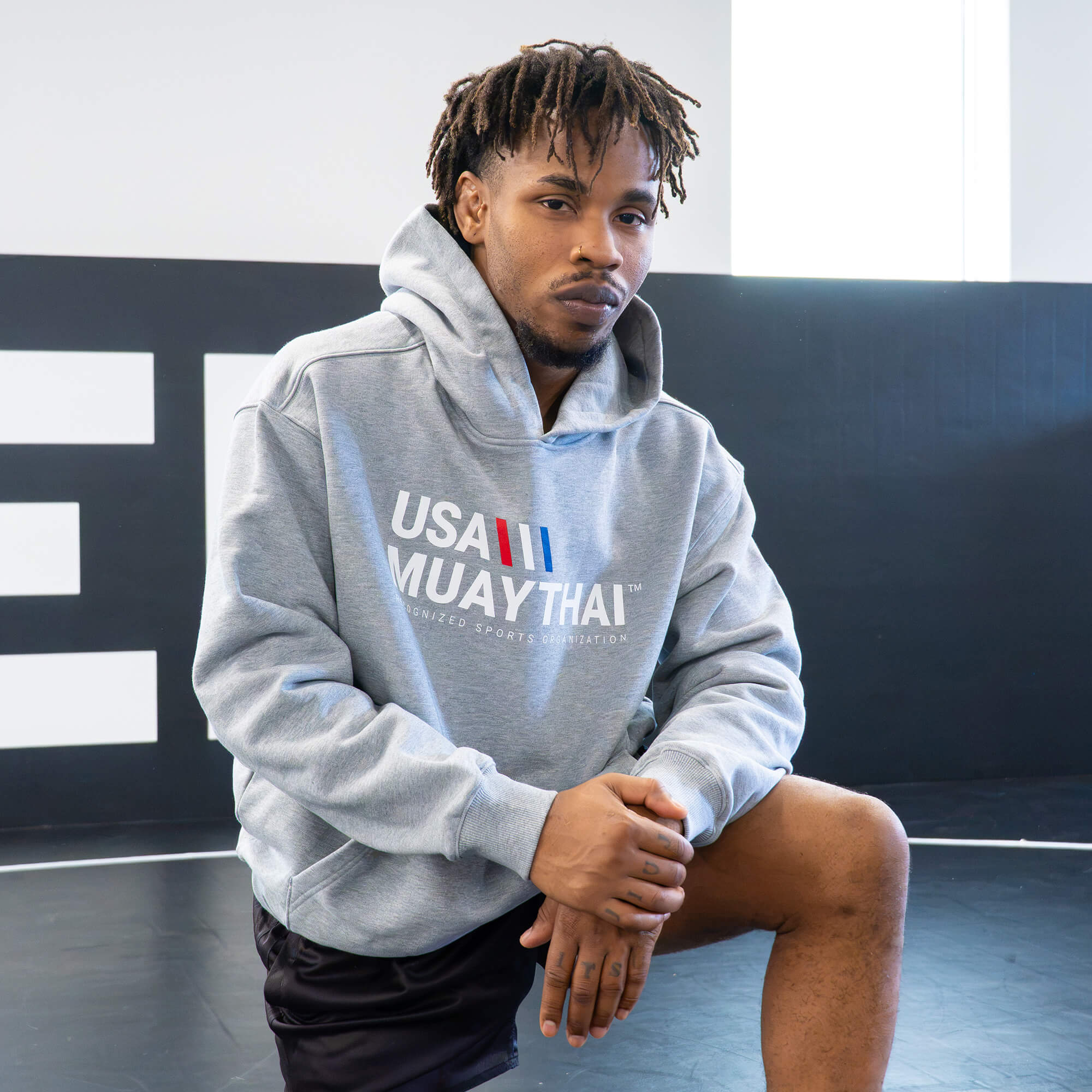 A male athlete kneeling in a gym while wearing the gray Combat Corner USA Muay Thai lifestyle hoodie, demonstrating its athletic, relaxed fit.