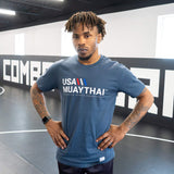 A male athlete standing with hands on his hips inside a gym mat area, modeling the front of the dark slate blue Combat Corner USA Muay Thai lifestyle t-shirt to show its fit and chest graphics.