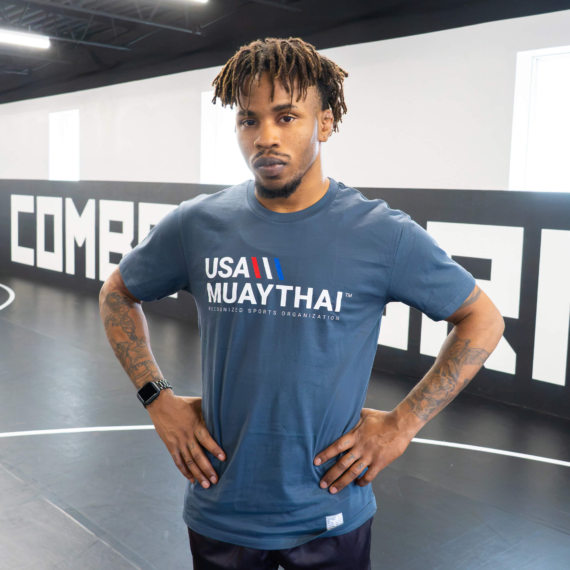 A male athlete standing with hands on his hips inside a gym mat area, modeling the front of the dark slate blue Combat Corner USA Muay Thai lifestyle t-shirt to show its fit and chest graphics.