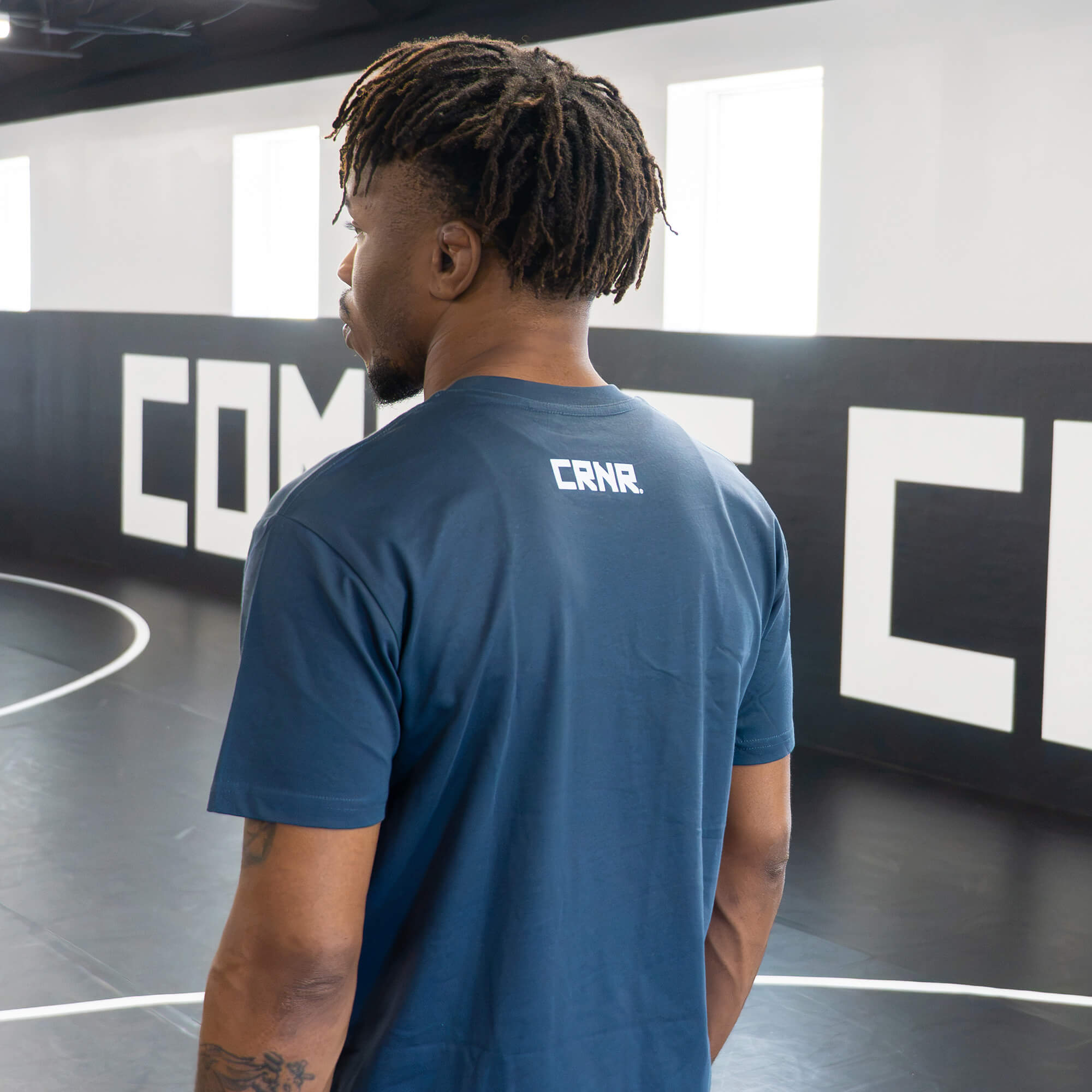 Over-the-shoulder view of a male athlete in a gym, modeling the back of the dark slate blue Combat Corner t-shirt to showcase the placement of the white CRNR logo on the upper back.