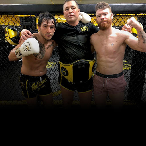 A trainer and two fighters posing together inside an MMA cage, with the trainer wearing a black long-sleeve rash guard and a yellow and black Combat Corner Belly Pad protector. 