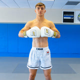 A front-facing studio portrait of a male fighter standing on grey gym mats in front of a blue wall. He is shirtless, wearing white Combat Corner hybrid fight shorts, and holding his white Combat Corner boxing gloves together across his chest.